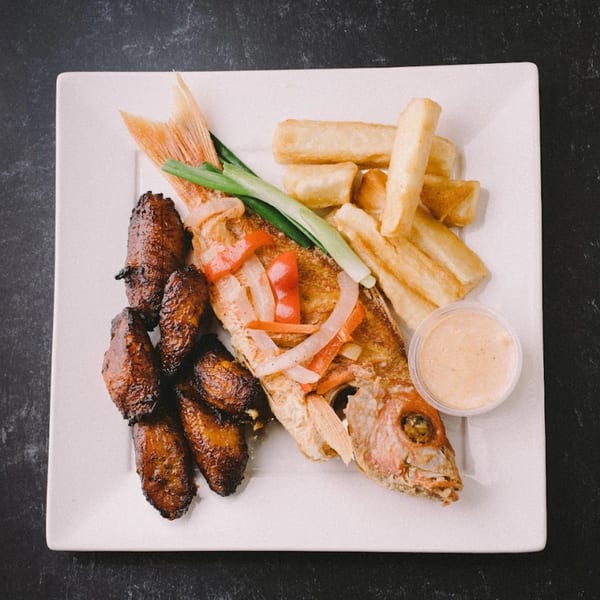 Whole fried fish topped with onions and peppers, served with plantains, fries, and sauce.