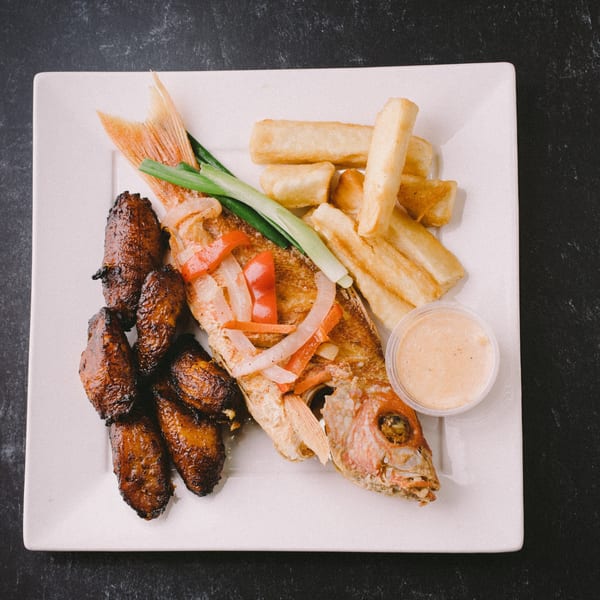 Whole fried snapper with plantains, yuca fries, and a dipping sauce on a white plate.