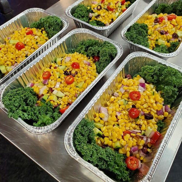 Multiple foil containers filled with vibrant corn salad, garnished with fresh kale, tomatoes, and olives.