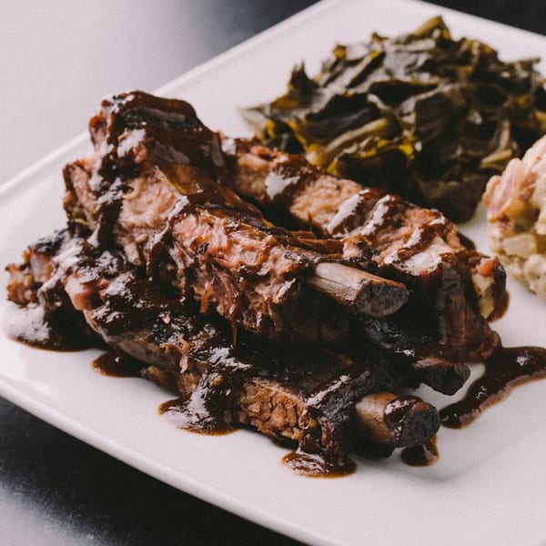 Jammin Island BBQ ribs with dark sauce, collard greens, and potato salad on a white plate.